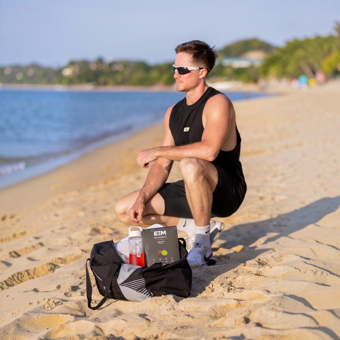 Man in the E3M Training Vest crouching on the beach beside an E3M Pre Workout box and gym bag